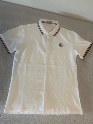 Vit pikétröja från Moncler - Klassisk vit pikétröja från Moncler med knappslå och broderad logga på bröstet. Kragen och ärmsluten har röd, vit och blå randig detalj. Perfekt till jeans eller shorts för en clean look.