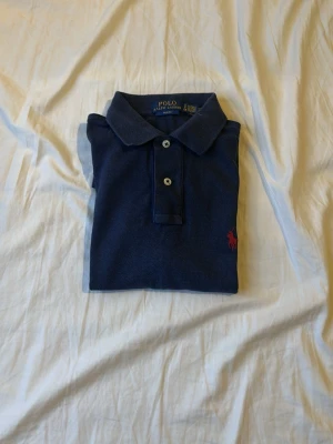 Marinblå pikétröja från Ralph Lauren - Klassisk marinblå pikétröja från Ralph Lauren med korta ärmar och krage. Tröjan har två knappar framtill och den ikoniska röda logotypen broderad på bröstet. Perfekt till jeans eller shorts för en clean och stilren look.