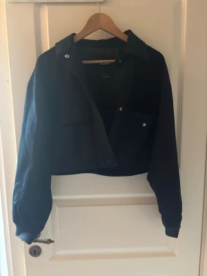 Mörkblå croppad jacka - Cool mörkblå croppad jacka med klassisk krage och knappar framtill. Jackan har en bröstficka med knapp och långa ärmar. Perfekt för att ge din outfit en edgy touch.