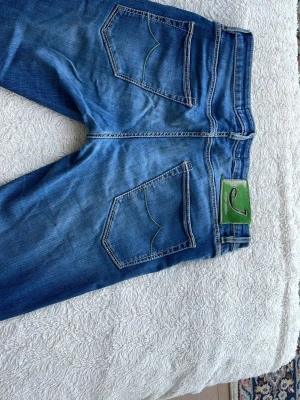 Blå jeans från Jacob Cohen - Snygga blå jeans från Jacob Cohen med gröna detaljer,  byxorna är använda och det förekommer en lagning på en byxbenet. Jeansen har normal midja, raka ben och är tillverkade i mjuk denim med tydlig tvättad effekt. Passar till uppklätt såsom skjorta och kavaj men även perfekta i mer vardagliga sammanhang!