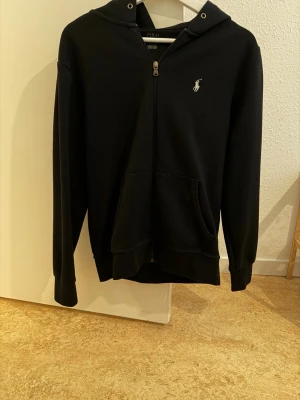 Svart hoodie från Polo Ralph Lauren - Snygg svart hoodie från Polo Ralph Lauren med dragkedja framtill och klassisk logga broderad på bröstet. Hoodien har huva, känguruficka och ribbade muddar vid ärmslut och nederkant. Tillverkad i mjukt bomullsmaterial för skön känsla.