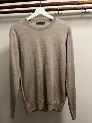 Beige stickad tröja Oscar Jacobson - Snygg beige stickad tröja från Oscar Jacobson med rund halsringning och ribbade muddar. Tillverkad i Italien och har en stilren, minimalistisk design som passar perfekt till många olika outfits.