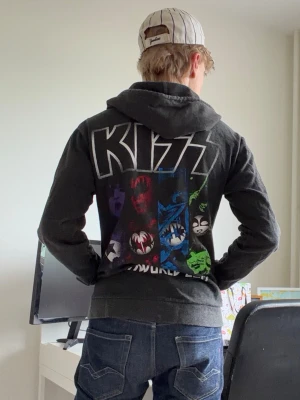 KISS zip hoodie med tryck (kissworld 2017) - Mörkgrå zip hoodie med stort färgglatt KISS-tryck på ryggen och mindre tryck med bandmedlemmarnas ansikten på bröstet. Huva med vita snören och dragkedja framtill. Perfekt för dig som gillar rock och vill sticka ut med en ikonisk bandtröja.
