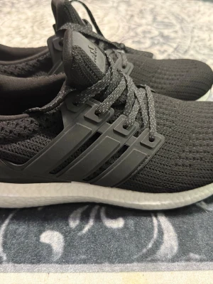 Svarta Adidas Ultra Boost sneakers - Svarta Adidas Ultra Boost sneakers med stickad ovandel och klassiska tre ränder i plast på sidan. Skorna har en vit Boost-sula för extra komfort och svarta skosnören med grå detaljer. Perfekta för en sportig och trendig look.