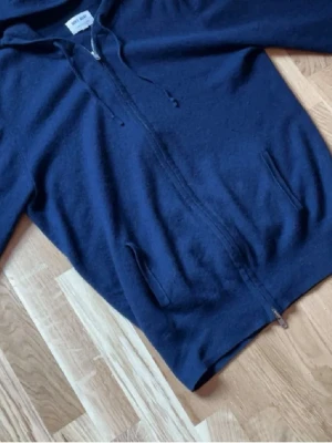 Mörkblå hoodie från Soft Goat - Mörkblå hoodie från Soft Goat i mjukt material, troligtvis kashmirblandning. Dragkedja framtill, två fickor och justerbar huva med snören. Perfekt för en chill och avslappnad stil. Litet hål vid bakre axeln men den syns knappt och går lätt att sy.