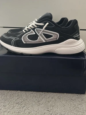 Svarta sneakers med vita detaljer - Snygga svarta sneakers med vita och grå detaljer samt vita skosnören. Skorna har en chunky sula i vitt och meshpaneler för extra komfort. Ikonisk logga på sidan och plös. Perfekta för dig som gillar streetwear och exklusiv stil. Använda en gång. Pris kan diskuteras. Storlek 41 men passar 42. Tveka inte att höra av dig!
