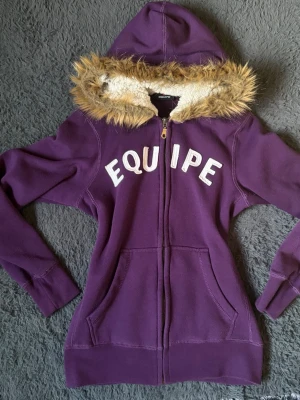 VINTAGE Lila hoodie från Equipe med fuskpäls - Lila hoodie från Equipe med dragkedja, stora fickor framtill och texten 'EQUIPE' i vitt på bröstet. Huva med mysigt vitt teddyfoder och kantad med fuskpäls. Perfekt för dig som vill ha en varm och snygg hoodie med unik look.