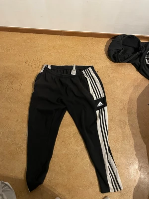 Svarta Adidas mjukisbyxor med ränder - Säljer ett par svarta mjukisbyxor från Adidas med klassiska vita ränder längs sidorna och Adidas-logga på benet. Byxorna har resår i midjan och är tillverkade i ett mjukt och bekvämt material, perfekta för chill eller träning.