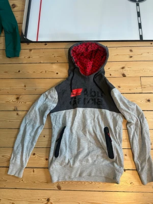 Grå hoodie från Abu Garcia - Snygg grå hoodie från Abu Garcia med svart och röd logga på bröstet. Huvan har röd insida med svart mönster och tröjan har svarta dragkedjor på fickorna. Perfekt för dig som gillar streetwear och vill ha något unikt.