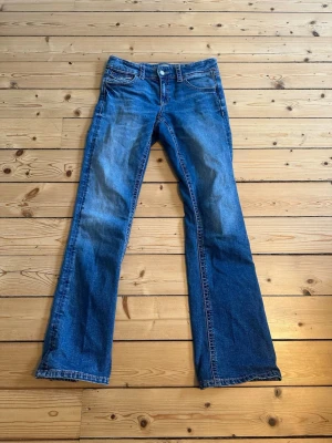 Blå bootcut jeans med slitningar - Säljer ett par blå bootcut jeans med snygga slitningar och klassisk femficksdesign. Jeansen har låg midja och är tillverkade i denim med en lätt tvättad look. Perfekta för en avslappnad och trendig stil.
