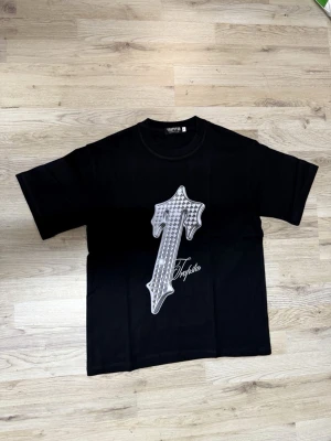 Svart T-shirt från Trapstar - Svart T-shirt från Trapstar med stort grafiskt T i vitt och grått framtill. Klassisk rund hals och korta ärmar. Oversized passform för en avslappnad stil. Perfekt för dig som gillar streetwear och vill sticka ut med en unik design.