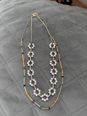 Dubbelhalsband med pärlor och färgglada pärlor - Snyggt dubbelhalsband med ett lager av vita pärlor formade som blommor och ett lager med små färgglada pärlor i orange, turkos, svart, lila och guld. Kedjan är i metall och halsbandet har en trendig boho-vibe. Perfekt för att lyfta din outfit med lite extra färg.