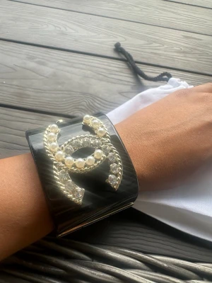 Chanel svart armband med pärlor - Säljer ett statement armband från Chanel i svart blank plast med bred design. Armbandet har en stor guldfärgad Chanel-logga dekorerad med vita pärlor och kristaller. Stängs med guldfärgat spänne och har märkning på insidan.