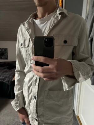 Beige overshirt - Stilren beige overshirt med klassisk krage och svarta knappar framtill. Två bröstfickor med lock och knapp samt två sidofickor. Tillverkad i bomull, perfekt för lager på lager och en avslappnad look.