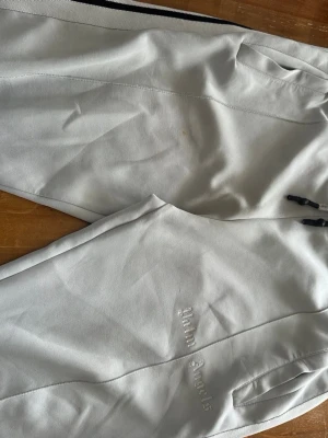 Palm Angels ljusgrå mjukisbyxor - Palm Angels mjukisbyxor i ljusgrått med broderad logga på benet. Byxorna har dragkedjor vid bensluten och svarta/vita ränder längs sidorna. Sköna och stilrena, perfekta för en chill look. Materialet känns mjukt och följsamt.