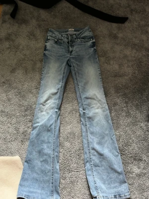 Ljusblå bootcut jeans med fickdetaljer - Säljer ett par ljusblå bootcut jeans med slitningar och snygga fickdetaljer bak. Jeansen har klassisk femficksmodell, låg midja och vida ben. Perfekta för dig som gillar retrostil och vill ha ett par jeans som sticker ut lite extra.