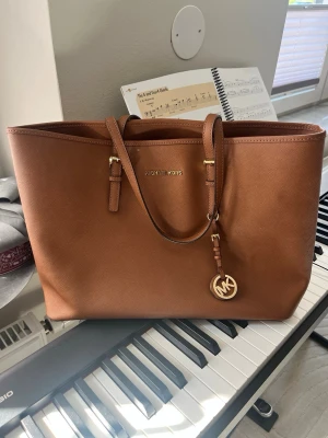 Brun väska från Michael Kors - Snygg brun handväska från Michael Kors i klassisk tote-modell. Väskan är gjord i slätt läder med guldfärgade detaljer och har ett hängande MK-emblem. Perfekt storlek för att få plats med allt du behöver och har dubbla axelband.