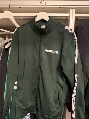 Askari - Sportig mörkgrön zip-tröja från Askari med vit logga på bröstet och breda svarta och vita detaljer längs ärmarna. Tröjan har hög krage, två fickor och ribbade muddar. Perfekt för en avslappnad och cool look.