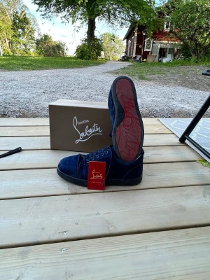 Blåa Christian Louboutin sneakers - Snygga blåa sneakers från Christian Louboutin med klassisk röd sula och svart kant. Skorna är tillverkade i mocka och har högt skaft. Perfekta för dig som vill sticka ut med exklusiva detaljer och ikonisk design.