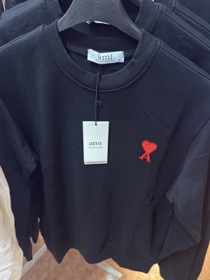Svart sweatshirt från Ami Paris - Svart sweatshirt från Ami Paris med rund halsringning och ett rött hjärtemblem broderat på bröstet. Tillverkad i mjuk bomull och har långa ärmar samt ribbade muddar vid ärmslut och nederkant. Enkel och stilren design som passar till det mesta.
