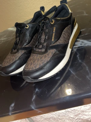 Michael Kors bruna och svarta sneakers - Snygga sneakers från Michael Kors i brunt och svart med gulddetaljer. Skorna har ett klassiskt MK-mönster på tyget, svarta läderdetaljer och vita sulor med guldfärgade inslag på hälen. Perfekta för dig som vill ha en trendig och lyxig look.