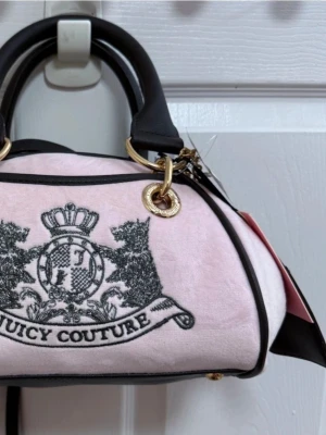 Rosa handväska från Juicy Couture - Söt rosa handväska från Juicy Couture i mjukt tyg med svarta detaljer och guldfärgade metalldelar. Väskan har ett broderat emblem framtill, svarta handtag och en dekorativ tag med krona samt ett gnistrande hjärthänge.