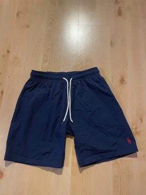 Marinblå shorts från Polo Ralph Lauren - Snygga marinblå shorts från Polo Ralph Lauren med vit snörning i midjan och klassisk röd logga broderad på benet. Perfekta för sommaren och har en avslappnad passform. Tillverkade i ett lätt syntetmaterial som torkar snabbt.