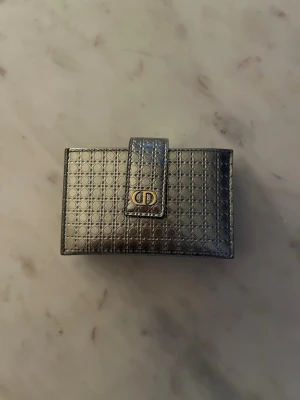 Dior plånbok - Snygg silverfärgad plånbok från Dior i glansigt läder med ikoniskt mönster och guldfärgad CD-logga på knäppet. Plånboken har flera fack för kort och sedlar samt en praktisk tryckknappsstängning. Perfekt accessoar för dig som gillar lyxiga detaljer.
