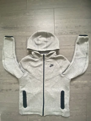 Nike tech  - Snygg ljusgrå hoodie från Nike med svart logga på bröstet. Hoodien har hel dragkedja framtill, två svarta fickor och en stor huva. Tillverkad i mjukt och bekvämt material, perfekt för en sportig och avslappnad stil.