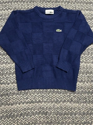 Blå stickad tröja från Lacoste - Mörkblå stickad tröja från Lacoste med rutmönster i stickningen och den klassiska gröna krokodilloggan på bröstet. Tröjan har rund halsringning, ribbade muddar och nederkant. Perfekt för dig som gillar stilrena och sportiga plagg. Den är i barn medium!