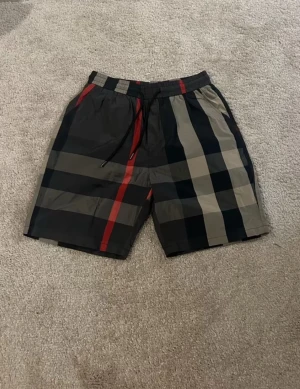 Burbery shorts  - Snygga rutiga shorts från Burberry med klassiskt mönster i svart, beige och rött. Shortsen har elastisk midja med snörning och är tillverkade i ett lätt och bekvämt material, perfekta för sommardagar. Shortsten passar L. Priset kan diskuteras vid snabbäffar