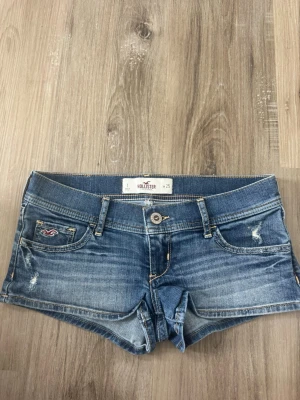 Blå denimshorts från Hollister - Säljer ett par klassiska blå denimshorts från Hollister med låg midja och slitna detaljer. Shortsen har fem fickor, bälteshällor och knappgylf. Perfekta för varma dagar och en avslappnad look.