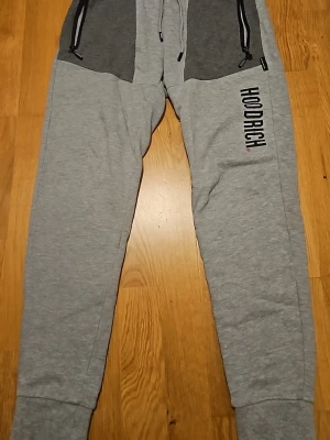 Grå mjukisbyxor från Hoodrich - Snygga grå mjukisbyxor från Hoodrich med svarta detaljer och dragkedjefickor framtill. Byxorna har en relaxed fit och resår vid bensluten. Tillverkade i mjukt bomullsmaterial, perfekta för chill dagar. Hoodrich-logga tryckt längs benet för extra streetkänsla. Säljer de för slutade använda där man bytte stil inget fel med de nästa som nya