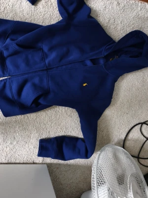 Blå hoodie från Polo Ralph Lauren - Snygg blå hoodie från Polo Ralph Lauren med dragkedja framtill och klassisk gul logga broderad på bröstet. Tillverkad i mjuk bomull och har en huva samt fickor på sidorna. Perfekt för en chill och stilren look.