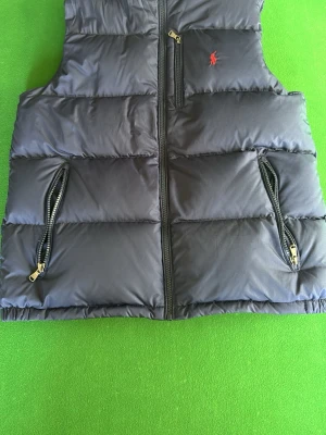dunväst från Polo Ralph Lauren - Snygg blå Ralph lauren väst. Västen är barn XL och sitter som en S. Bra skick (bara att höra av sig om fler bilder) säljer då jag inte använder den. Västen är köpt på Åhléns i Stockholm så kan garantera att den är äkta