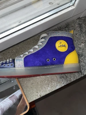 Christian Louboutin höga sneakers - Unika höga sneakers från Christian Louboutin i blå mocka med silverglittrande detaljer, gul häl och logga på sidan. Tåpartiet är täckt av silverfärgade nitar och sulan är röd undertill. Skorna har vita skosnören och en cool mix av material och färger.