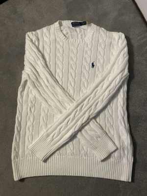 Vit kabelstickad tröja Polo Ralph Lauren - Kabelstickad vit tröja från Polo Ralph Lauren med rund halsringning och den klassiska blå logotypen broderad på bröstet. Använd ett väldigt få tal gånger. Perfekt för en clean och stilren look. Hör av er med frågor👍🤩