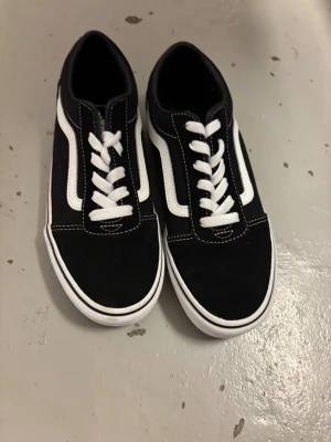 Svarta Vans Old Skool sneakers - Klassiska Vans Old Skool sneakers i svart med vit sula och vit sidostripes. Skorna har låga skaft, vita snören och är tillverkade i en kombination av mocka och canvas. Perfekta för en streetwear-look och passar till det mesta.
