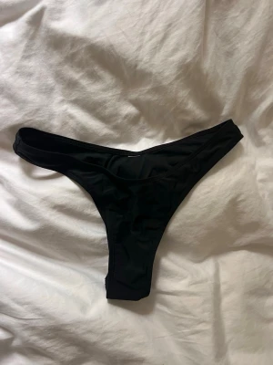 Svart bikiniunderdel i tanga-modell (endast testad) - Säljer en  svarta bikiniunderdel i tanga-modell med hög skärning, oklart vart den är köpt då lapp är bortklippt (kanske Shein). Klassisk och stilren design i stretchigt material som passar perfekt till stranden eller poolen. Inga synliga loggor eller detaljer. Materialet känns mjukt och följsamt. Endast testad. Gissar på strl S.