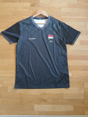Svart Irak fotbollströja Suwar - Snygg svart fotbollströja från Suwar med Irak-emblem på bröstet och diskret mönster över hela framsidan. Tröjan har korta ärmar, V-ringning med grå kant och tryckt logga. Perfekt för dig som vill stötta Irak på match eller träning.