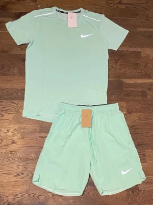 Nike miller - Snyggt mintgrönt träningsset från Nike med matchande t-shirt och shorts. Båda plaggen har Nike-logga i vitt och är tillverkade i ett lätt och snabbtorkande material, perfekt för träning eller sportaktiviteter. T-shirten har rund hals och shortsen har resår i midjan. Priset kan diskuteras eftersom de fick lite färg i sig som går bort i tvätt.