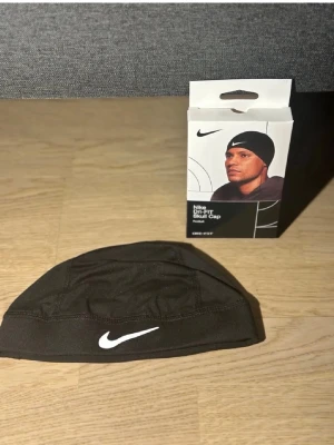 Nike svart Dri-FIT skull cap - Nike Dri-FIT skull cap i svart funktionsmaterial med vit logga framtill. Perfekt för träning eller under hjälm, tillverkad i stretchigt och snabbtorkande tyg som håller svetten borta. Snygg och sportig design med låg profil.