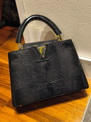 Lizard Capucines - black lizard-embossed finish with gold-tone hardware. A timeless and sophisticated design inspired by one of Paris' most iconic fashion houses. The glossy texture catches the light beautifully and gives the bag an exclusive look. Perfect for everyday wear, dinners, events, or whenever you want to elevate an outfit. ✨ Features: • Black lizard-embossed finish • Gold-tone hardware • Structured top-handle design • Removable & adjustable shoulder strap included • Dustbag included • Multiple interior 