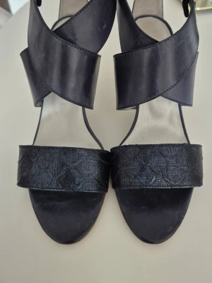 Svarta höga sandaletter med korsade remmar - Snygga blå sandaletter med höga klackar och breda korsade remmar i läder. Främre remmen har ett diskret mönster som ger extra stil. Öppen tå och elegant design som passar perfekt till sommarens outfits.