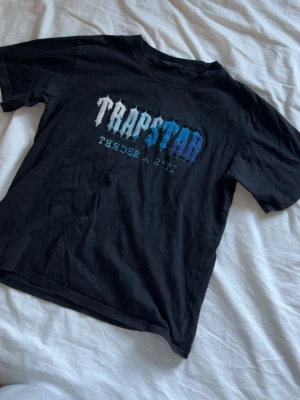Svart Trapstar t-shirt med blå text - Trapstar t-shirt i svart med tryckt logga i vitt och blått på bröstet. Klassisk rund hals och korta ärmar. Tillverkad i mjuk bomull för skön känsla och avslappnad passform. Perfekt för dig som gillar streetwear och vill sticka ut.