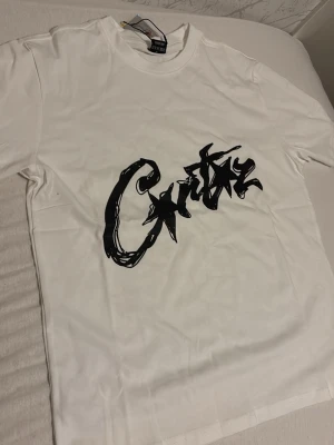 Vit t-shirt med svart graffiti-tryck - Säljer en vit t-shirt från Crutz med ett stort svart graffiti-inspirerat tryck på bröstet. Klassisk rund hals och korta ärmar. Perfekt för dig som gillar streetwear och vill sticka ut med en unik design.