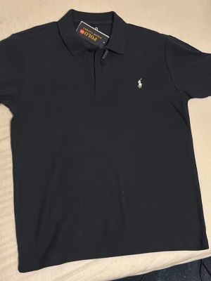 Mörkblå pikétröja från Polo Ralph Lauren - Klassisk svart pikétröja från Polo Ralph Lauren med vit broderad logga på bröstet. Tröjan har krage, knappar framtill och är tillverkad i mjuk bomull. Perfekt för en clean och stilren look.