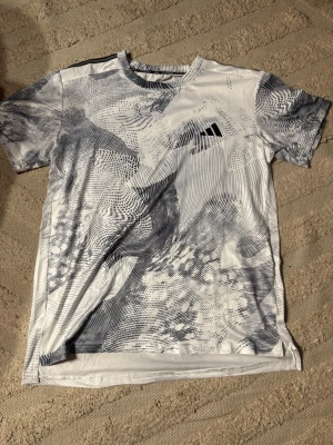 Vit Adidas Adizero t-shirt med mönster - Säljer en vit t-shirt från Adidas Adizero-kollektionen i storlek M/L med abstrakt grått mönster över hela tröjan. T-shirten har rund hals, korta ärmar och är tillverkad i ett lätt, tekniskt material som andas. Adidas-logga på bröstet och Adizero-tryck i nacken. Kom pm vid frågor!