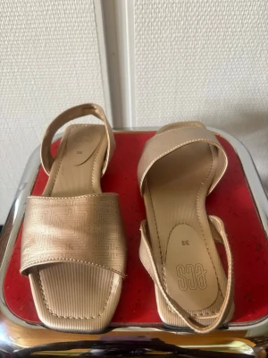 Guldiga sandaler med fyrkantig tå - Snygga sandaler i guldig färg från ECS med fyrkantig tå och bred rem framtill. Sandalerna har en diskret mönstrad yta och en smal hälrem. Perfekta för dig som vill ha en stilren och trendig look till sommaren.