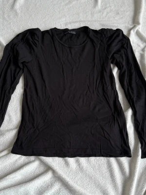 Svart långärmad topp från Zara (S) - Stilren svart långärmad topp från Zara med puffiga axlar och rund halsringning. Tillverkad i mjuk bomull som känns skön mot huden. Perfekt att matcha med jeans eller kjol för en trendig look.